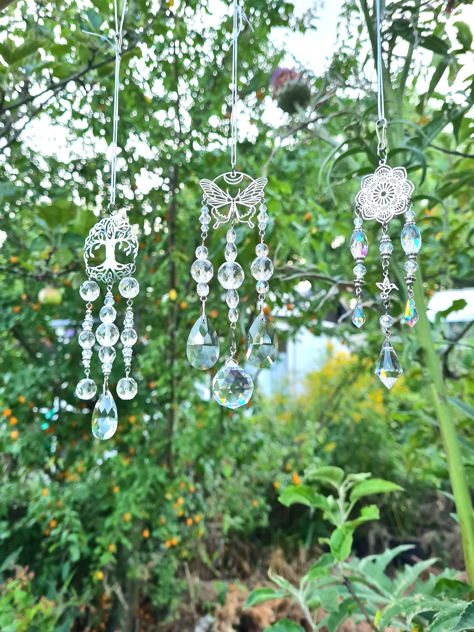 Drie handgemaakte kristallen suncatchers met levensboom, vlinder en mandala, hangend tussen het groen